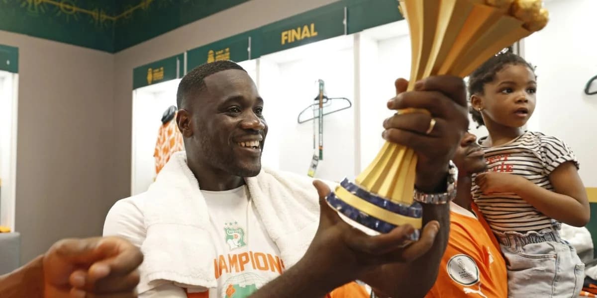 Emerse Faé souriant, tenant un ballon de football, entouré de supporters.